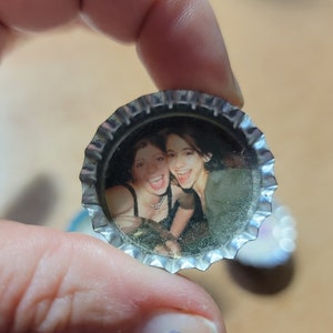 May include: A bottle cap with a printed image of two smiling people. The cap is silver with a crimped edge, and the photo inside shows two women with dark hair. The cap is held between fingers.