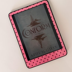 May include: A pink e-reader case with black polka dots surrounds a digital screen displaying the book cover of "Conform" by Ariel Sullivan. The cover art features a stylized tower and clouds.