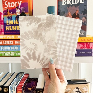 Op de afbeelding: Twee decoratieve kaarten, een met een wit en grijs tie-dye patroon en de andere met een wit en grijs ruitjespatroon. De kaarten worden in een hand gehouden.