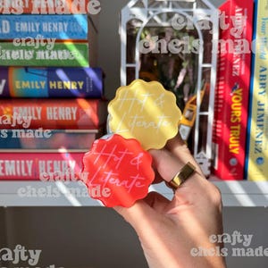 Peut inclure: Une étagère remplie de livres colorés, dont les titres incluent "Happy Place" et "Beach Read". Deux objets en forme de fleur avec le texte "Hot & Literate" sont tenus devant les livres. Une cage à oiseaux décorative blanche est également visible.