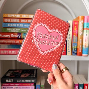 Puede incluir: Funda de teléfono de lunares rojos y blancos con un corazón rosa que dice "Fictional Characters". La funda se sostiene frente a una estantería llena de libros. La funda es una cubierta protectora transparente.
