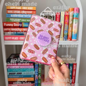 May include: A pink phone case with a pattern of hot dogs and white stars, featuring a purple pop socket that reads "DO NOT DISTURB." The case is held in front of a white bookshelf filled with colorful books.