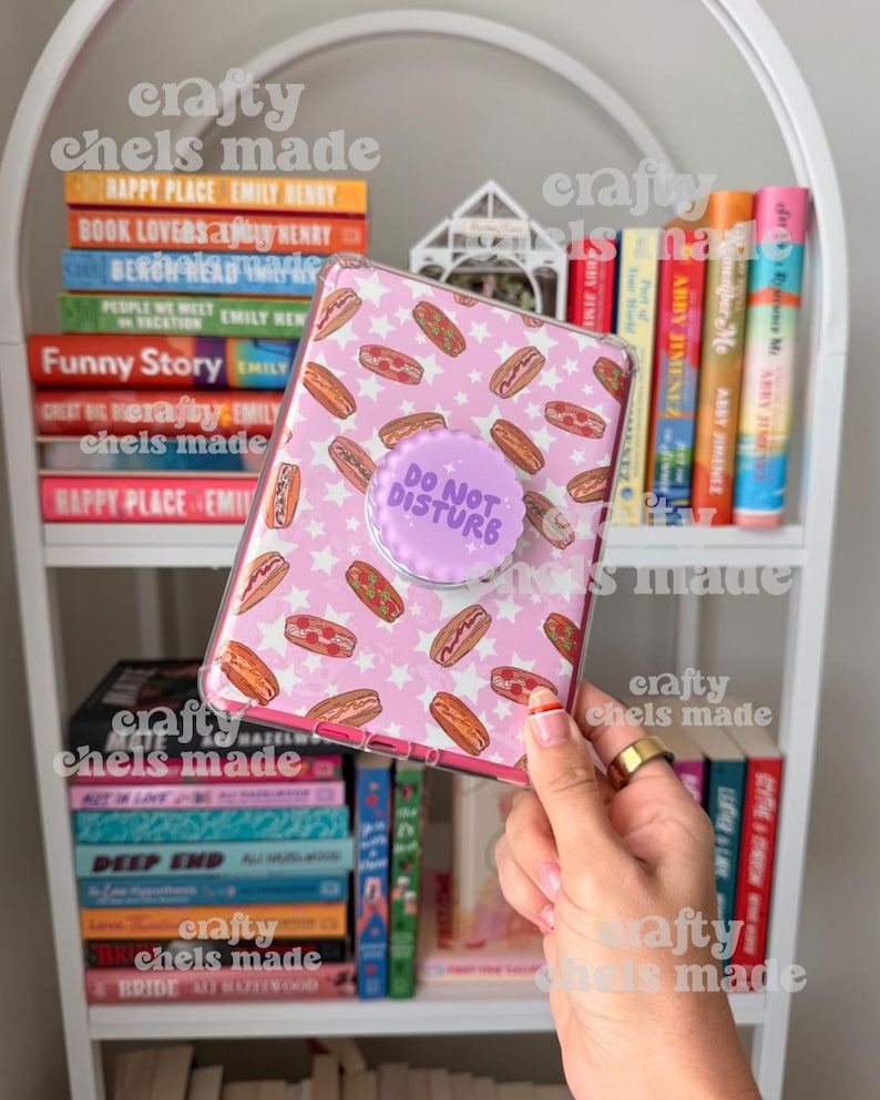 May include: A pink phone case with a pattern of hot dogs and white stars, featuring a purple pop socket that reads "DO NOT DISTURB." The case is held in front of a white bookshelf filled with colorful books.