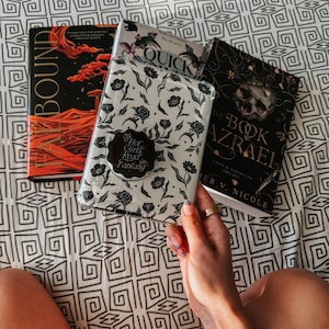 May include: A white phone case with a floral design and a black sticker that says "Hot Girls Read Fantasy". The case is being held by a hand with a ring on the finger. There are three books in the background: one with a red and orange cover that says "Bound", one with a blue and white cover that says "Quick", and one with a black cover that says "The Book of Azrael".