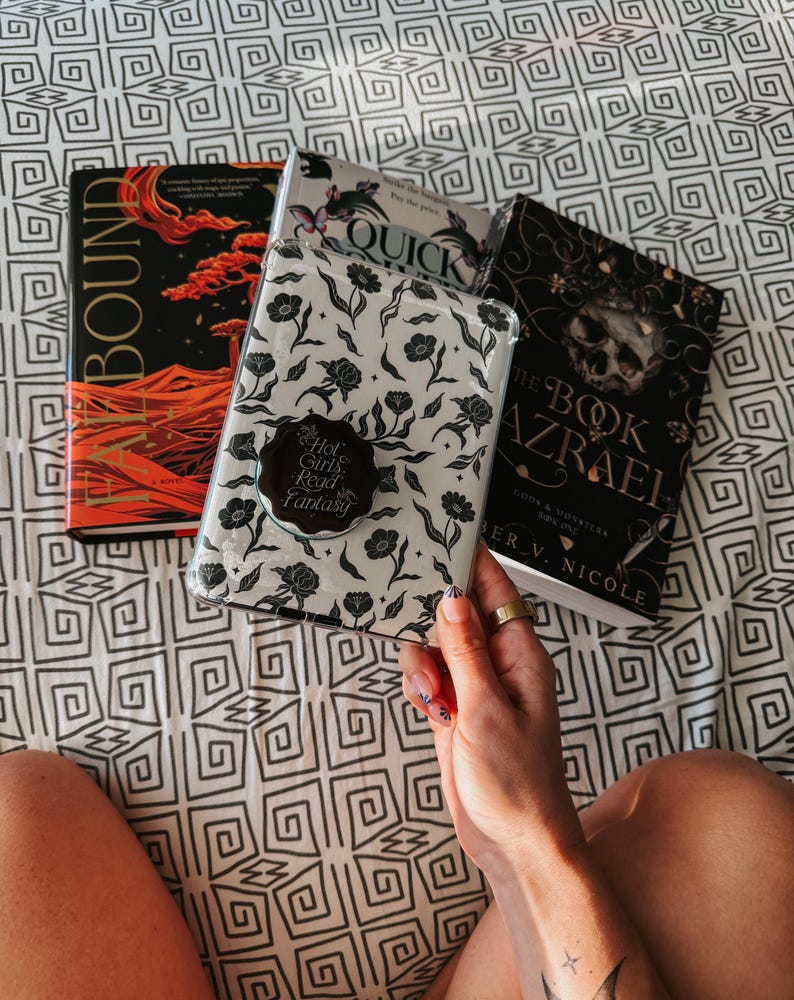 May include: A white phone case with a floral design and a black sticker that says "Hot Girls Read Fantasy". The case is being held by a hand with a ring on the finger. There are three books in the background: one with a red and orange cover that says "Bound", one with a blue and white cover that says "Quick", and one with a black cover that says "The Book of Azrael".