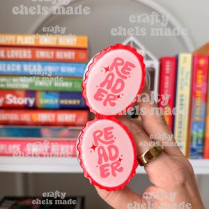 May include: Two red and white bottle cap magnets with the word "READER" in pink script. The magnets are held in front of a bookshelf filled with colorful books. The text "crafty chels made" is visible in the image.