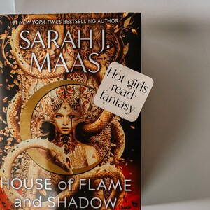 May include: A hardcover book titled "House of Flame and Shadow" by Sarah J. Maas. The cover features a woman with snakes and a gold crescent moon design. A sticker reads "Hot girls read+ fantasy."