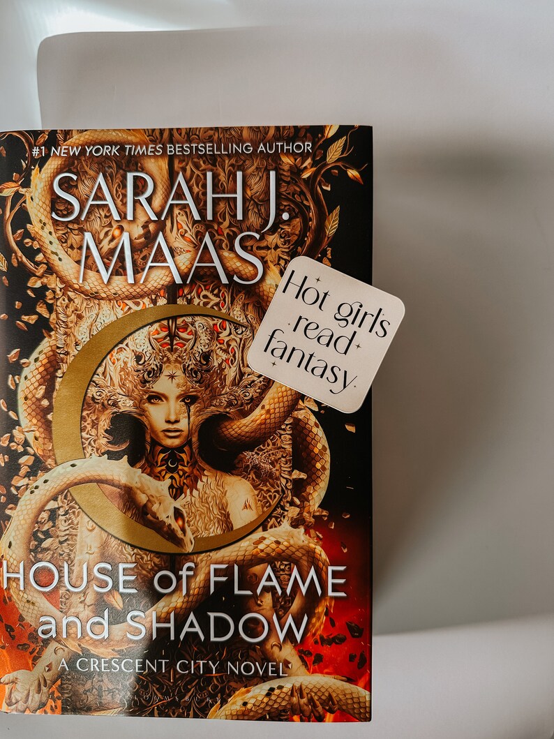 May include: A hardcover book titled "House of Flame and Shadow" by Sarah J. Maas. The cover features a woman with snakes and a gold crescent moon design. A sticker reads "Hot girls read+ fantasy."