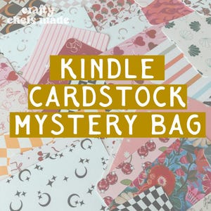 May include: A collection of patterned cardstock papers in various designs, including cherries, stripes, and floral patterns. The text "KINDLE CARDSTOCK MYSTERY BAG" is displayed in a gold rectangle. The papers are in shades of pink, white, and black.