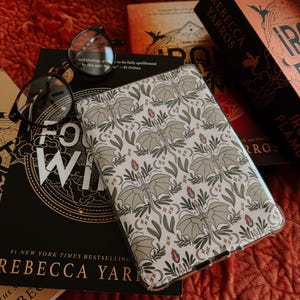 May include: A clear phone case with a white background and a repeating pattern of green leaves and bats. The case is on top of a stack of books with the title "REBECCA YARROS" visible.
