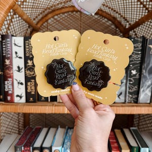 May include: Two yellow cards with black text that reads "Hot Girls Read Fantasy" and "Kindle Grip". The cards have a black wax seal with the same text. The cards are held in front of a bookshelf with many books.