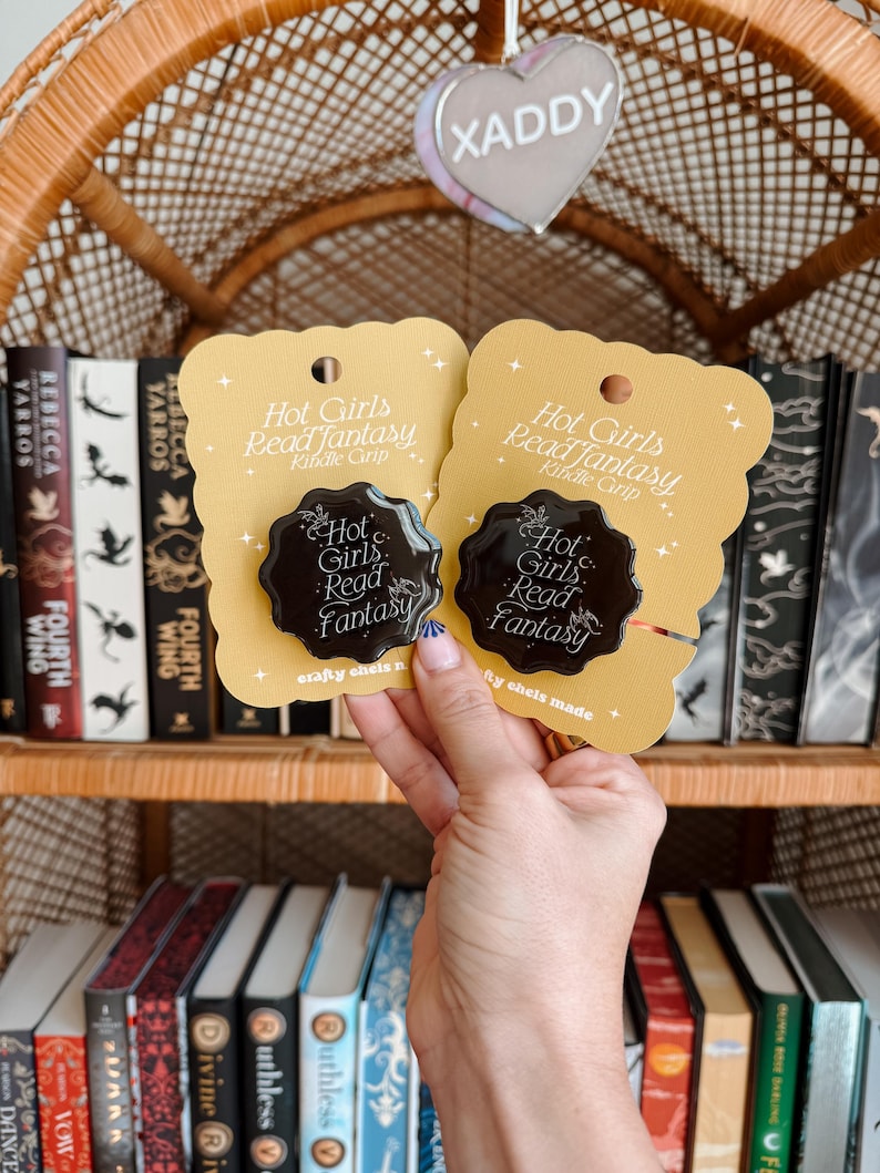 May include: Two yellow cards with black text that reads "Hot Girls Read Fantasy" and "Kindle Grip". The cards have a black wax seal with the same text. The cards are held in front of a bookshelf with many books.