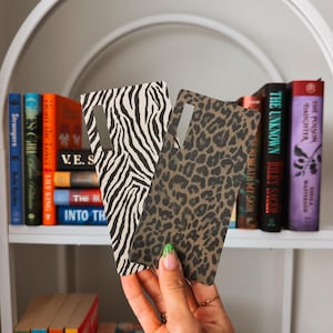 May include: Two rectangular phone cases featuring distinct animal prints. One case displays a black and white zebra stripe pattern, while the other showcases a brown and black leopard spot design. Both cases include a vertical cutout for a camera lens.
