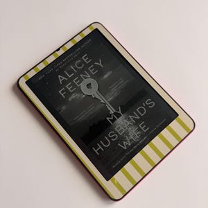 May include: An e-reader with a pink border and a cover featuring the book "My Husband's Wife" by Alice Feeney. The cover has a key graphic and a black and white image. The e-reader has a striped design.