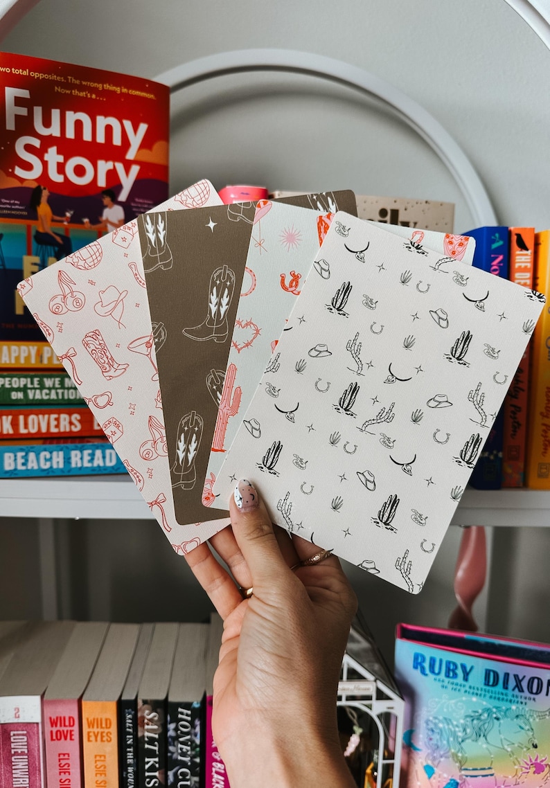 May include: Four different patterned paper notebooks with a western theme. The patterns include cacti, cowboy hats, horseshoes, and cowboy boots. The notebooks are held in a hand.