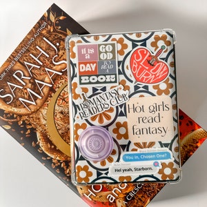 May include: An open book and a tablet case with stickers that read "It is a good day to read a book", "Romantasy Readers Club", and "Hot girls read fantasy". A pop socket is attached to the case.