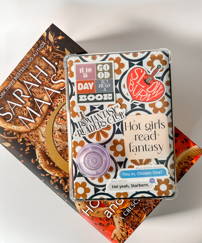 May include: An open book and a tablet case with stickers that read "It is a good day to read a book", "Romantasy Readers Club", and "Hot girls read fantasy". A pop socket is attached to the case.