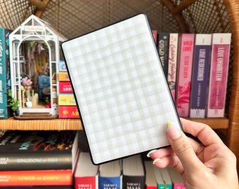 Puede incluir: Una cubierta de libro a cuadros blancos y grises con un borde negro. La cubierta está siendo sostenida por una mano frente a una estantería llena de libros.