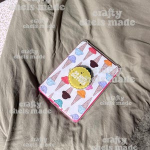 May include: A clear phone case with a white background featuring colorful ice cream cones. A yellow pop socket with the text "DO NOT DISTURB" is attached. The case is on a wrinkled, olive-green fabric.