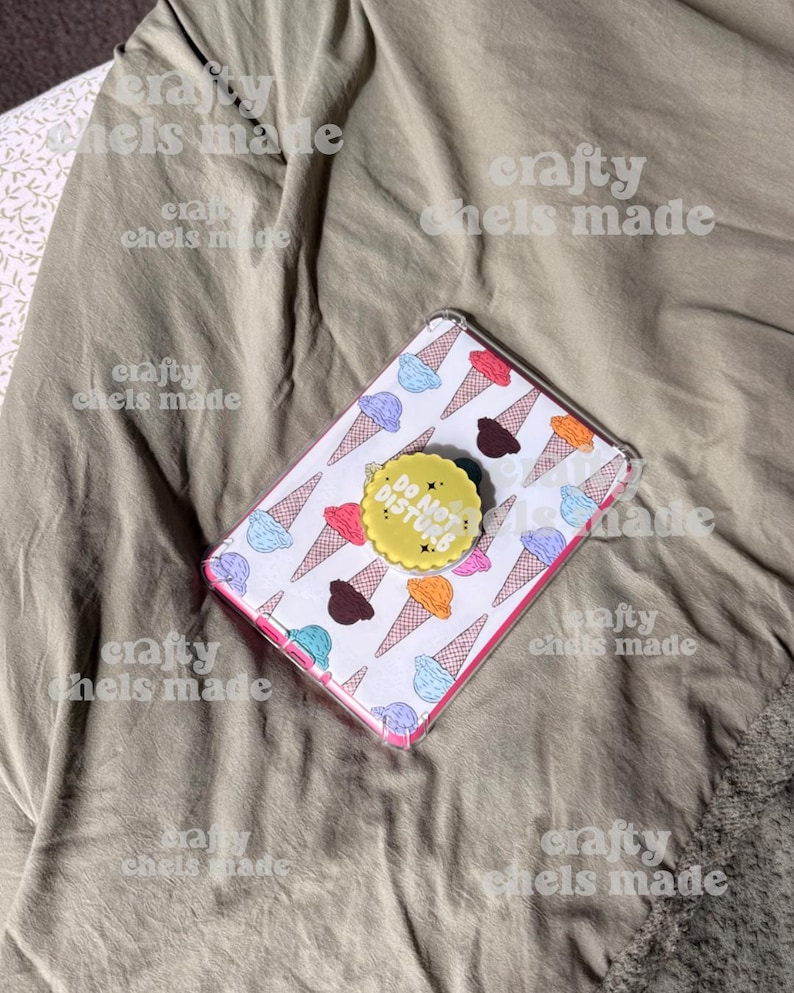 May include: A clear phone case with a white background featuring colorful ice cream cones. A yellow pop socket with the text "DO NOT DISTURB" is attached. The case is on a wrinkled, olive-green fabric.