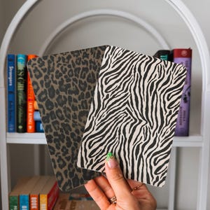May include: Two rectangular book sleeves featuring animal prints. One sleeve displays a brown and black leopard spot pattern, while the other features a black and white zebra stripe design. Both items have rounded corners and a textured surface finish.