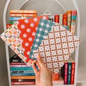 May include: A hand holds a stack of patterned square cards in front of a white bookshelf filled with books. The cards feature geometric and starburst designs in various colors, including red, blue, and tan. The bookshelf has books with titles like "Happy Place" and "Book Lovers".