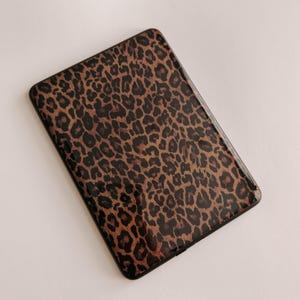 May include: A rectangular electronic tablet or e-reader device featuring a protective cover with a brown and black leopard print pattern. The device has rounded corners and a smooth, glossy finish across the entire front surface area.