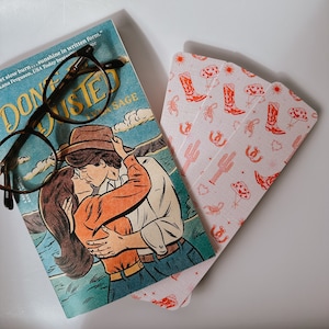 May include: A book with a couple embracing on the cover, with the title "Don't be Dissed" and the text "FRAM ARE RAN NOVEL". The book is on top of a stack of pink and white bookmarks with a cowboy boot and scorpion design.