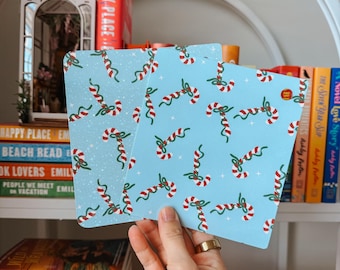 Könnte beinhalten: Zwei hellblaue quadratische Karten mit einem sich wiederholenden Muster aus rot-weißen Zuckerstangen und grünen Bändern. Die Karten werden vor einem Bücherregal mit Büchern gehalten. Die Karten haben ein festliches, weihnachtliches Thema.