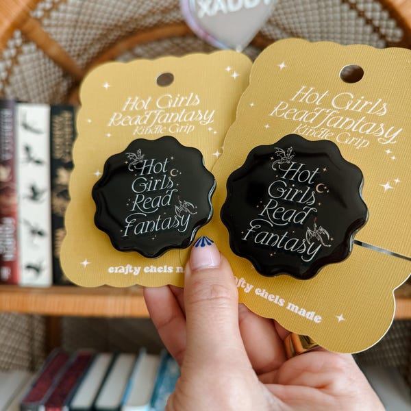 Puede incluir: Dos pines de esmalte negro y dorado con el texto "Hot Girls Read Fantasy" y un pequeño diseño de dragón. Los pines están sobre un fondo amarillo con estrellas blancas.