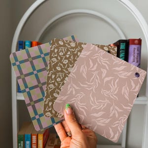May include: Three square swatches with rounded corners, each featuring a distinct pattern. One has a green and purple plaid design, another displays a white floral print on a brown background, and the third shows a white leaf pattern on a mauve base.