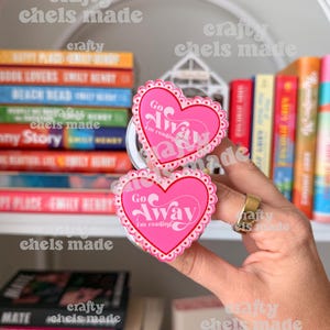 May include: Two pink heart-shaped phone grips with the text "Go Away I'm reading" are held in front of a bookshelf. The grips have a white scalloped border. Books with colorful spines are visible.