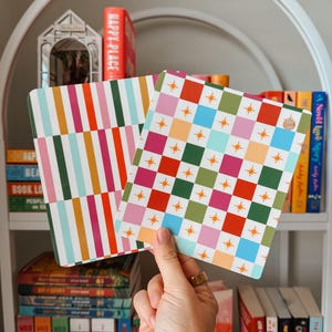 May include: Two decorative square coasters with colourful patterns are held up in front of a bookshelf. One coaster has vertical stripes in various colours, and the other has a checkerboard design with stars. The bookshelf is filled with books.