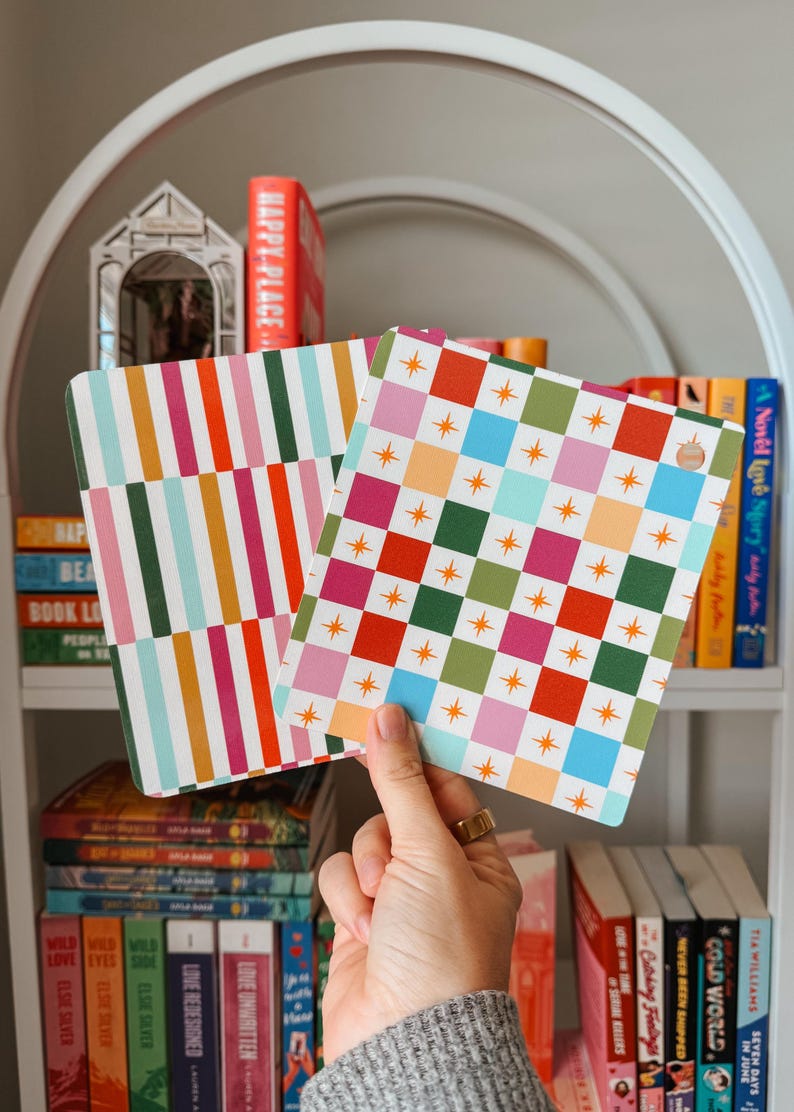 May include: Two decorative square coasters with colourful patterns are held up in front of a bookshelf. One coaster has vertical stripes in various colours, and the other has a checkerboard design with stars. The bookshelf is filled with books.