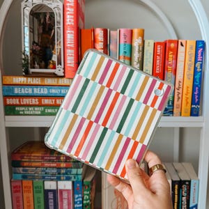 May include: A clear tablet case with a colourful striped pattern in pink, orange, green, and blue is held in front of a white bookshelf filled with books. The books have titles like "Happy Place" and "Beach Read."