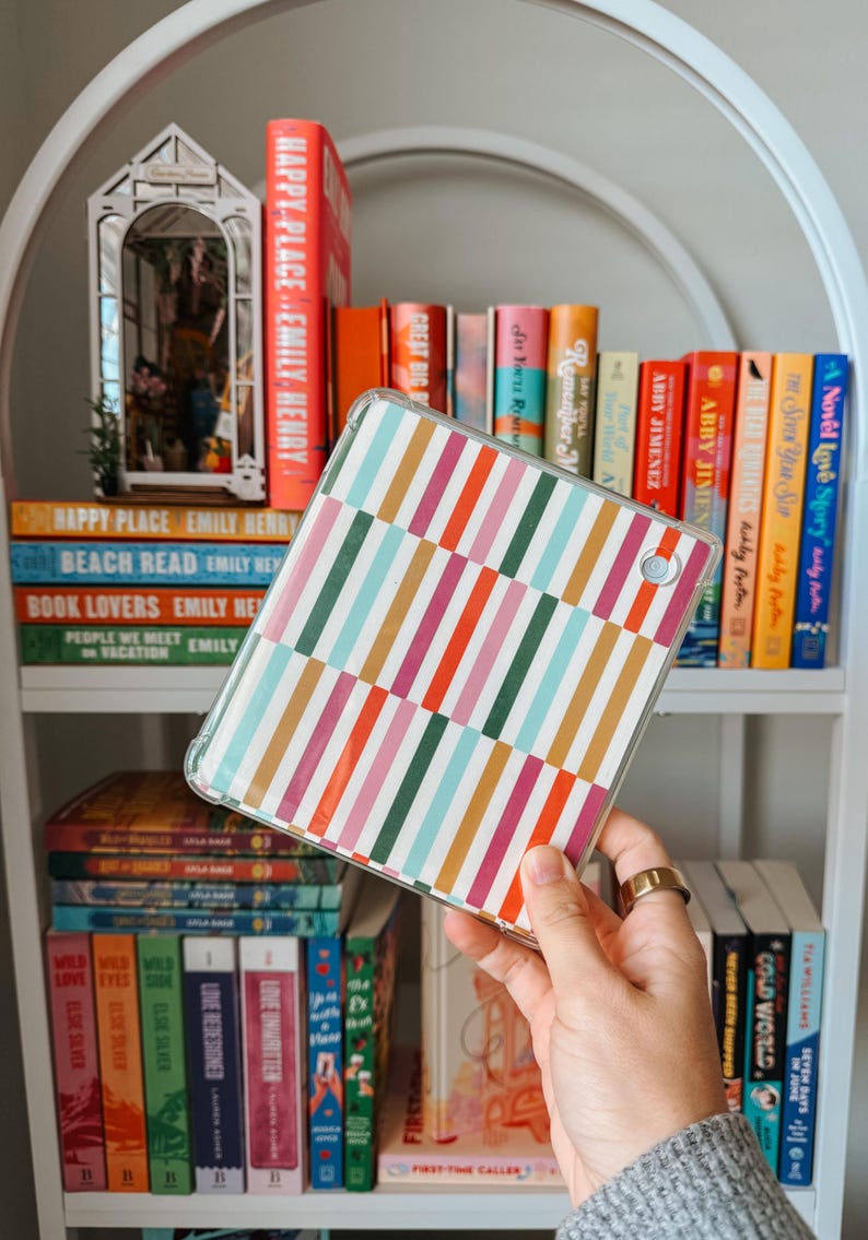 May include: A clear tablet case with a colourful striped pattern in pink, orange, green, and blue is held in front of a white bookshelf filled with books. The books have titles like "Happy Place" and "Beach Read."