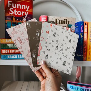 May include: Four different patterned notebooks with a western theme. The notebooks feature designs of cacti, cowboy hats, horseshoes, and cowboy boots. The notebooks are stacked on top of each other and are being held by a hand.