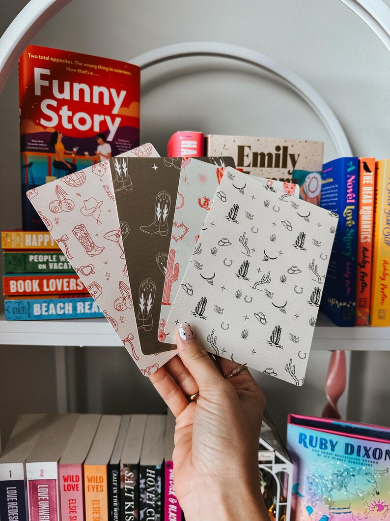 May include: Four different patterned notebooks with a western theme. The notebooks feature designs of cacti, cowboy hats, horseshoes, and cowboy boots. The notebooks are stacked on top of each other and are being held by a hand.