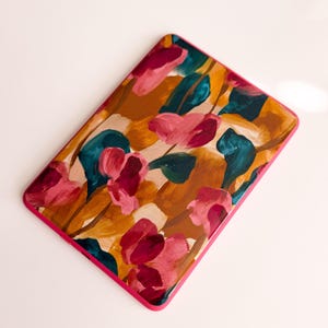 May include: A rectangular electronic reader cover featuring a vibrant painted floral pattern with shades of magenta, deep teal, and golden ochre on a light beige background, finished with a solid bright pink border around the edges.