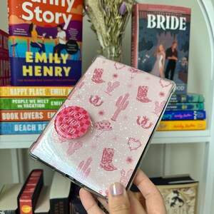 May include: A pink and white phone case with a glittery finish and a pink cowboy boot pattern. The case has a pink and white button that says "Hot Girls Read".