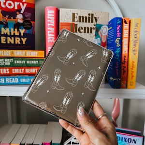 May include: A clear phone case with a brown background and a white pattern of cowboy boots. The case is being held in a hand with white and black nail polish.