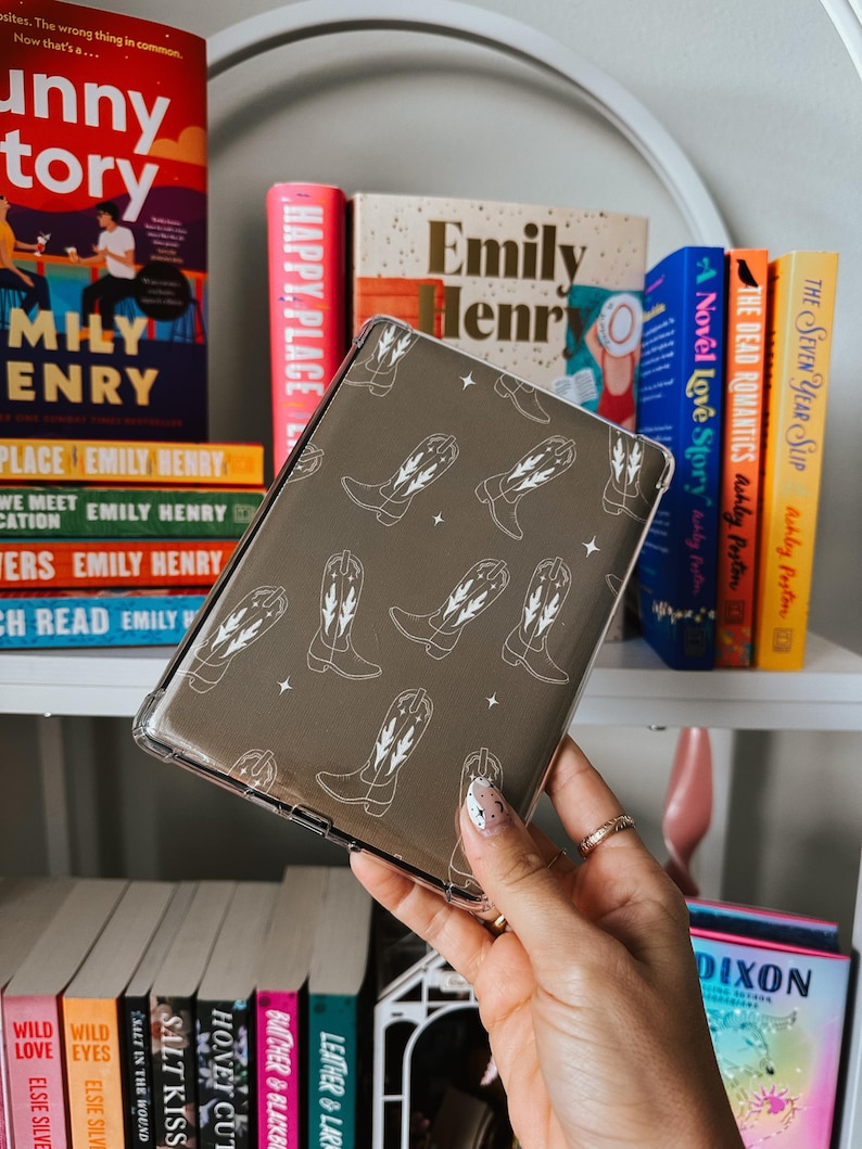 May include: A clear phone case with a brown background and a white pattern of cowboy boots. The case is being held in a hand with white and black nail polish.