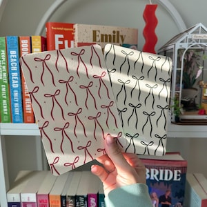 May include: Two small notebooks with a white background and a repeating pattern of red or black bows. The notebooks are held in a hand against a bookshelf with books.