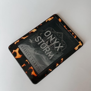 May include: A black e-reader with a tortoiseshell case. The screen displays the cover of the book "Onyx Storm" by Rebecca Yarros.
