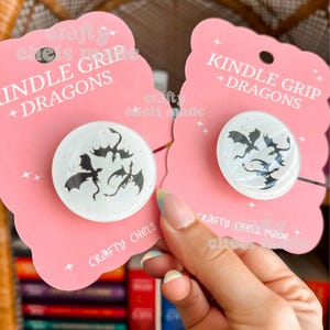 Könnte beinhalten: Zwei weiße Kindle-Griffe mit schwarzen Drachensilhouetten auf rosa Karten. Die Karten tragen die Aufschrift "KINDLE GRIP + DRAGONS" und "CRAFTY CHELS MADE". Die Griffe werden von einer Person gehalten.