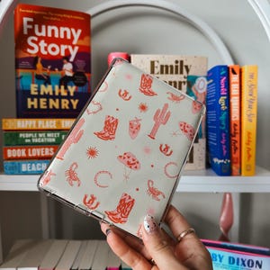 May include: A clear phone case with a light green background and a repeating pattern of red cowboy boots, cacti, scorpions, and stars. The case is being held in a hand.