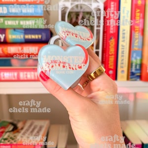 May include: Two heart-shaped book lockets with the text "MONSTER ROMANCE BOOK CLUB" in red and white on a light blue background. The lockets are held in front of a bookshelf filled with books.