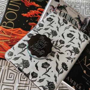 May include: A white phone case with a black floral pattern. The case has a black and gold sticker that says "Hot Girls Read Fantasy".