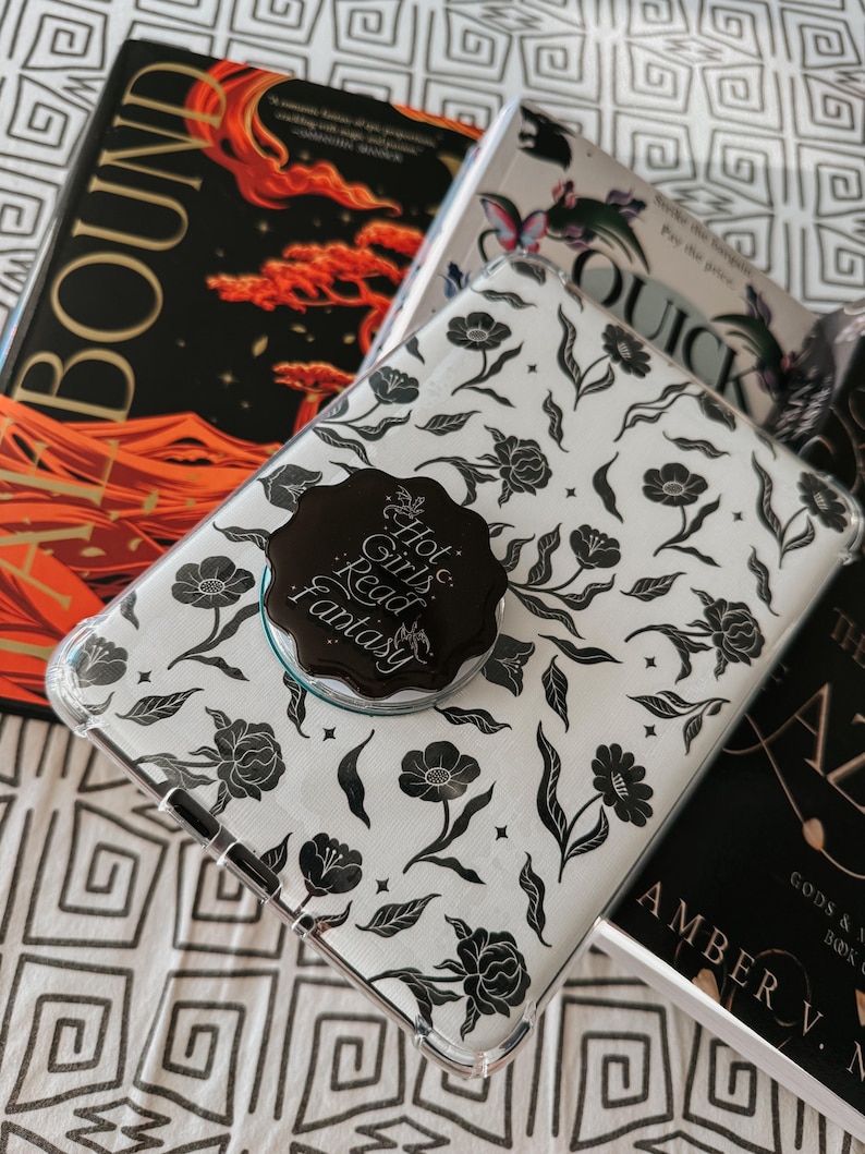 May include: A white phone case with a black floral pattern. The case has a black and gold sticker that says "Hot Girls Read Fantasy".