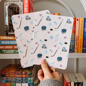 May include: Two rectangular cards with a white background and a repeating pattern of hockey skates, helmets, hockey sticks, pucks, snowflakes, hearts, and ribbons. The cards are held in front of a bookshelf.